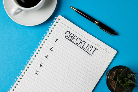 Spiral notebook with a handwritten numbered checklist beside a pen, black coffee and a potted succulent on a blue desk