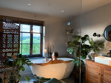 Guest relaxing in a freestanding bath in a boutique holiday let bathroom with carved wooden screens, houseplants, a wooden vanity and countryside views